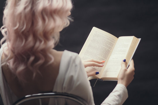 blonde girl holding open book sitting on a chair, back ofyoung woman reading a novel, concept of hobby, relaxation, leisure on black background