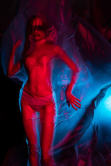 Slim girl dressed in underwear emotionally posing, wrapped in fluttering in the wind and fitting her graceful young body cellophane film, in blue and red light. Artistic, creative, abstract design