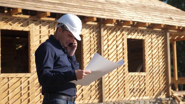 concept building constructing architect slow motion video. man builder in a helmet stands at a lifestyle construction holding a scheme house plan. site near a wooden frame house under construction