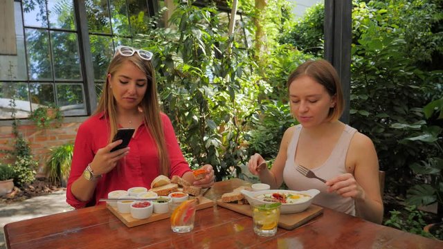 Two young women friends eat breakfast together take food photo of dishes for social media