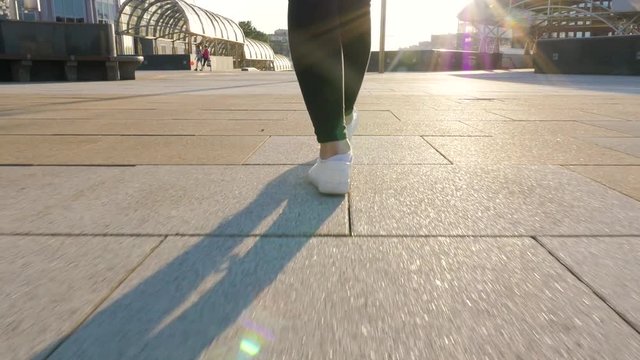 female legs in black trousers and white shoes walk along a clean, empty path. towards the sun, shooting from a low point, the camera follows the movement. city walk in slow motion close up