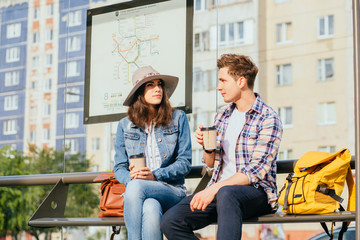 Handsome guy trying to get acquainted with pretty girl at bus stop. Man and woman travelers with backpacks drinking coffee, talking while sits on bench of bus station.