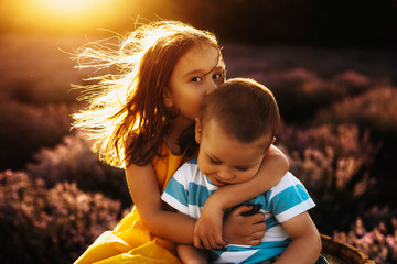 Portrait of a amazing little girl dressed in yellow dress embracing and kissing her little brother on his head while looking at camera against amazing sunset.