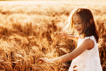 Portrait of a lovely little girl dressed in white dress touching wheat against sunset smiling.