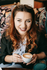 Close up portrait of a cheerful plus size caucasian woman with red hair drinking a coffee in a coffee shop and looking at camera smiling.
