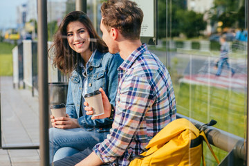 Positive relax couple traveler drinking coffee outdoor at stop while waiting tram, sits on bench at...