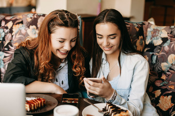 Portrait of two cheerful girlfriend looking to a smartphone screen smiling while sitting in a cafe eating cheesecake and drinking coffee.