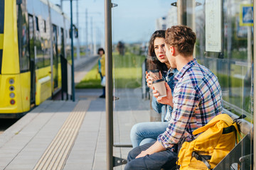 Positive relax couple traveler drinking coffee outdoor at stop while waiting tram, sits on bench at...