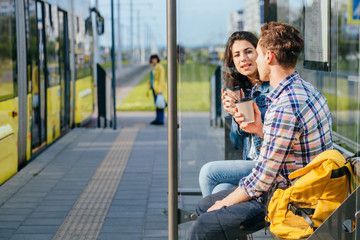 Positive relax couple traveler drinking coffee outdoor at stop while waiting tram, sits on bench at...