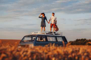Young couple man with a guitar and woman in a hat are standing on the roof of a car in a wheat...