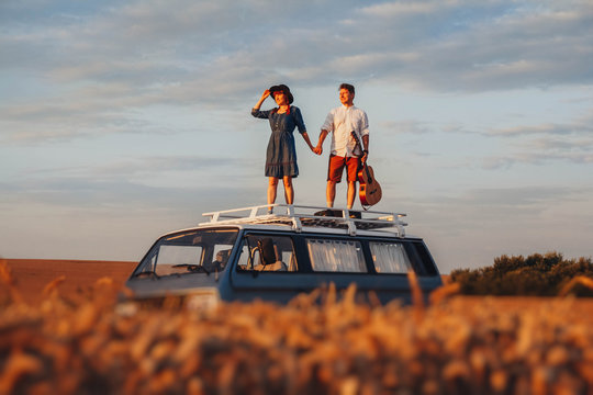 Young Couple Man With A Guitar And Woman In A Hat Are Standing On The Roof Of A Car In A Wheat Field. Travel And Adventur