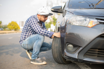 Fototapeta na wymiar Young man changing wheel on the car at the side of the road, young businessman has problems with the wheel of car. He is kneeing and looking at it with seriousness , Transportation, traveling concept