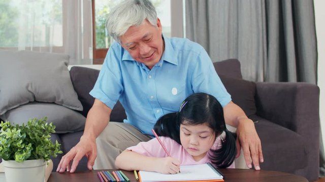 Asian grandfather teach granddaughter drawing and doing homework at home. Senior Chinese, grandpa happy relax with young girl lying on sofa in living room at home concept. Slow motion.