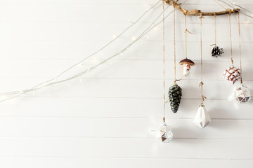 Christmas vintage toys hanging on wooden boho branch on background of white wall in festive modern room. Stylish glass ornaments, holiday decorations, eco style. Space for text