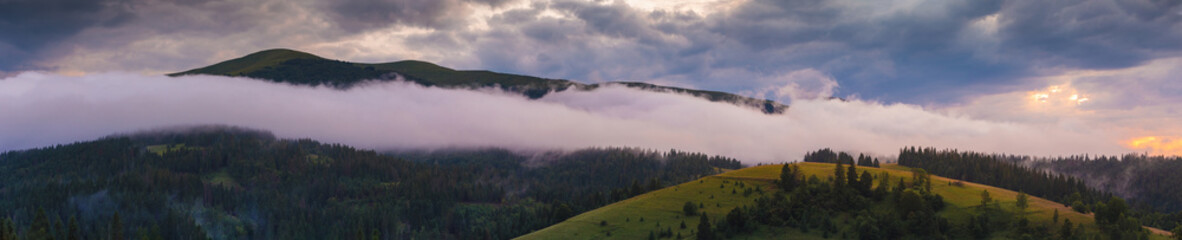Panorama of foggy mountains after the rain at sunset moment. Dreamy beautiful clouds over mountains slopes, covered with spruce forest. Carpathian mountains. Ukraine.