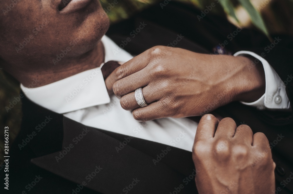 Poster closeup of an african-american male wearing a ring and a black suit jacket fixing his necktie