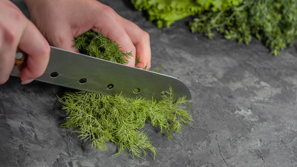Female hand cuts a juicy green bunch of dill a large knife on a dark table