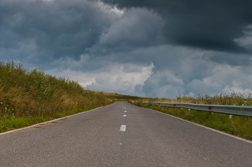 Empty rural asphalt road with green vegetation, dark dramatic gathering stormclouds on the sky.
