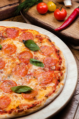 pepperoni pizza decorated with basil on a wooden board, the background is wooden decorated with vegetables and cutlery