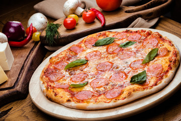 pepperoni pizza decorated with basil on a wooden board, the background is wooden decorated with vegetables and cutlery