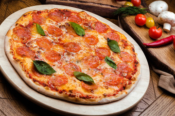 pepperoni pizza decorated with basil on a wooden board, the background is wooden decorated with vegetables and cutlery