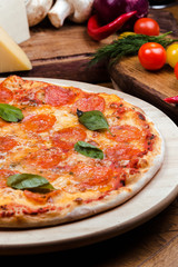 pepperoni pizza decorated with basil on a wooden board, the background is wooden decorated with vegetables and cutlery