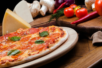 pepperoni pizza decorated with basil on a wooden board, the background is wooden decorated with vegetables and cutlery