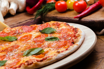 pepperoni pizza decorated with basil on a wooden board, the background is wooden decorated with vegetables and cutlery