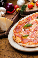 pepperoni pizza decorated with basil on a wooden board, the background is wooden decorated with vegetables and cutlery