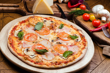 pizza with mushrooms, tomatoes, ham decorated with basil on a wooden board, the background is wooden decorated with vegetables and cutlery