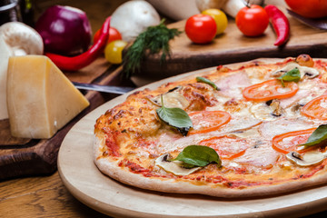 pizza with mushrooms, tomatoes, ham decorated with basil on a wooden board, the background is wooden decorated with vegetables and cutlery