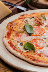 pizza with mushrooms, tomatoes, ham decorated with basil on a wooden board, the background is wooden decorated with vegetables and cutlery
