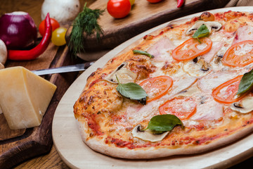 pizza with mushrooms, tomatoes, ham decorated with basil on a wooden board, the background is wooden decorated with vegetables and cutlery