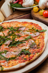 Italian pizza with ham, tomatoes, cheese and arugula on a wooden board, the background is wooden decorated with vegetables and cutlery