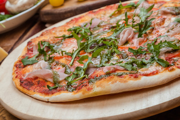 Italian pizza with ham, tomatoes, cheese and arugula on a wooden board, the background is wooden decorated with vegetables and cutlery