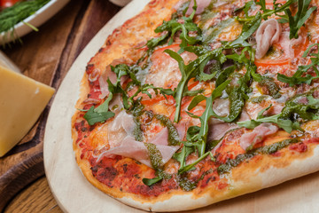 Italian pizza with ham, tomatoes, cheese and arugula on a wooden board, the background is wooden decorated with vegetables and cutlery
