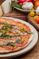 Italian pizza with ham, tomatoes, cheese and arugula on a wooden board, the background is wooden decorated with vegetables and cutlery
