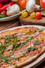 Italian pizza with ham, tomatoes, cheese and arugula on a wooden board, the background is wooden decorated with vegetables and cutlery