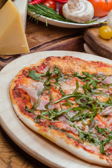Italian pizza with ham, tomatoes, cheese and arugula on a wooden board, the background is wooden decorated with vegetables and cutlery