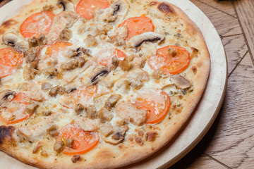 Italian pizza with chicken, tomatoes, mushrooms and cheese on a wooden board, the background is wooden decorated with vegetables and cutlery