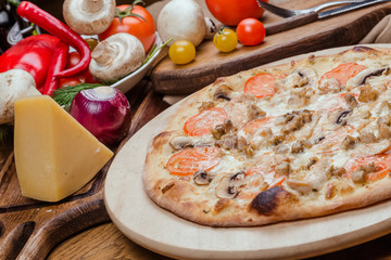 Italian pizza with chicken, tomatoes, mushrooms and cheese on a wooden board, the background is wooden decorated with vegetables and cutlery