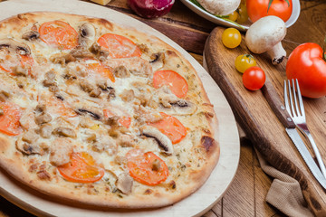 Italian pizza with chicken, tomatoes, mushrooms and cheese on a wooden board, the background is wooden decorated with vegetables and cutlery