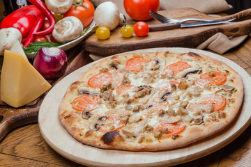 Italian pizza with chicken, tomatoes, mushrooms and cheese on a wooden board, the background is wooden decorated with vegetables and cutlery