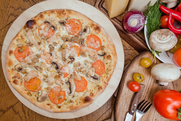 Italian pizza with chicken, tomatoes, mushrooms and cheese on a wooden board, the background is wooden decorated with vegetables and cutlery