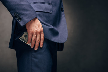 cropped view of businessman standing and putting dollar banknotes in pocket on grey