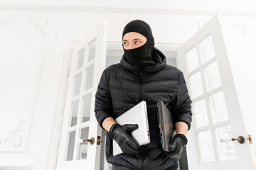 Thief with black balaclava stealing two modern expensive laptops. The burglar commits a crime in Luxury apartment with stucco.