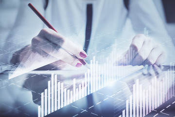 A woman hands writing information about stock market in notepad. Forex chart holograms in front. Concept of research.