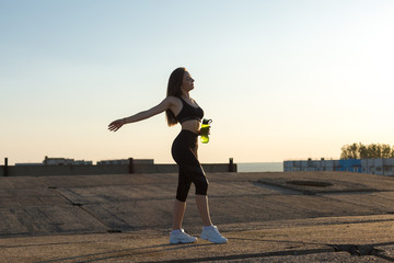 Slim athletic girl takes a break between classes and drinks water from a bottle, urban background. Beautiful sky at sunset.	