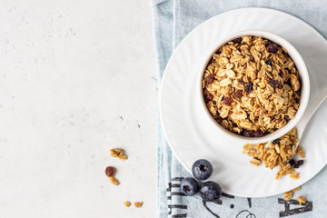 Homemade granola in a white ceramic bowl with raisins, fresh blueberries and milk. Healthy breakfast ingredients on light grey background.