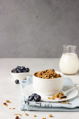 Homemade granola in a white ceramic bowl with raisins, fresh blueberries and milk. Healthy breakfast ingredients on light grey background.